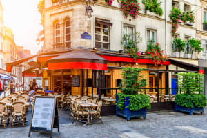 Parisian-style café terrace with wicker chairs, red awnings and leafy planters beneath balconies overflowing with flowers.