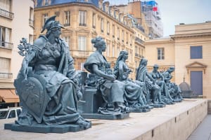Row of seated bronze female statues on stone plinths outside the Musée d’Orsay, with Parisian buildings behind them.