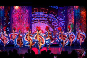 Moulin Rouge dancers perform an acrobatic French cancan routine in blue, white and red ruffled costumes.