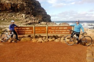 Photo of two guests with their bicycles standing on either side of the Cape of Good Hope sign-boards, at the nature reserve at Cape Point.