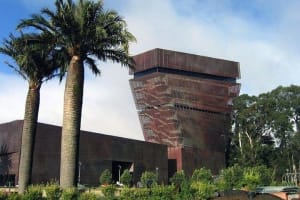 Modern brown brick building with dramatic tilted upper section, palm trees, and landscaping