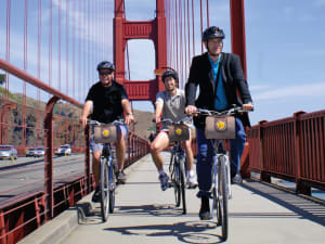 Three cyclists riding bikes on the Golden Gate Bridge pedestrian path with the iconic red suspension bridge structure in the background
