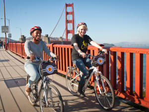 Two cyclists riding bikes on the Golden Gate Bridge pedestrian path in San Francisco with the iconic red suspension bridge and bay in background