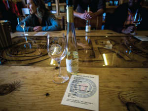 Wine tasting setup at a winery with a bottle, glass, and menu on a wooden table with guests in background