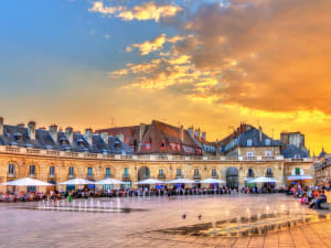 Wide view of Place de la Libération in Dijon at sunset with arcaded buildings, café terraces and water fountains reflecting the colorful evening sky