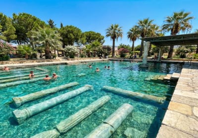 People swimming in Cleopatra's Antique Pool in Hierapolis, Pamukkale, with clear turquoise water and ancient Roman columns submerged under a bright blue sky.