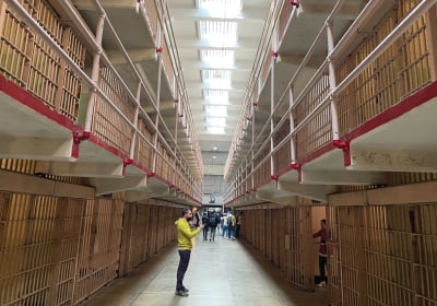 Interior of a historic prison with multiple tiers of wooden cell doors, metal railings, and visitors walking through the corridor.