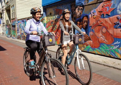 Two cyclists riding bikes with cargo baskets in front of colorful street mural art