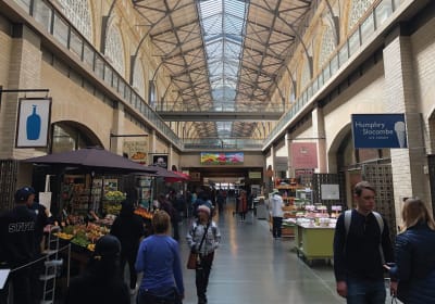 Interior of a bustling covered market hall with vendor stalls, shoppers, and a glass ceiling skylight