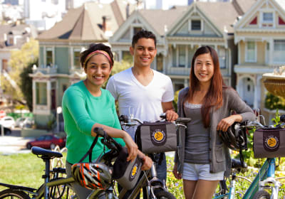 Three young adults with bikes and helmets smiling in front of Victorian houses in San Francisco