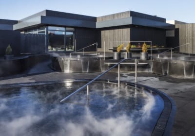 Exterior view of Krauma Geothermal Baths at dawn or dusk, showing multiple steaming circular pools set in dark stone terracing, with the ang