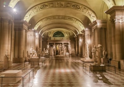 Marble statues displayed in a Louvre gallery with carved vaulted ceilings, tall columns and a patterned stone floor.