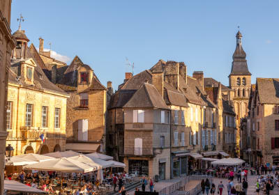 Busy square in Sarlat-la-Canéda with café terraces, golden-stone houses and the church tower in the background on a bright day