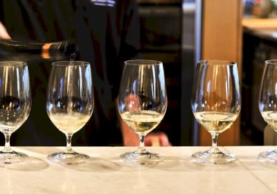 Line of wine glasses filled with white wine being poured at a tasting counter
