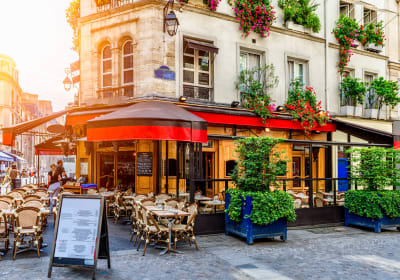 Parisian-style café terrace with wicker chairs, red awnings and leafy planters beneath balconies overflowing with flowers.