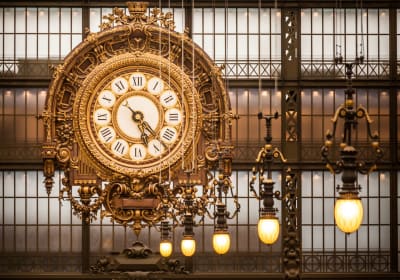 Ornate gilded clock with Roman numerals inside the Musée d’Orsay, beside glowing pendant lamps and tall gridded windows.