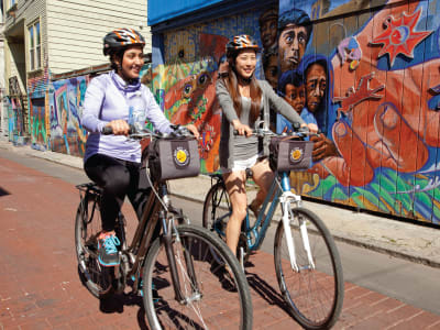 Two cyclists riding bikes with cargo baskets in front of colorful street mural art