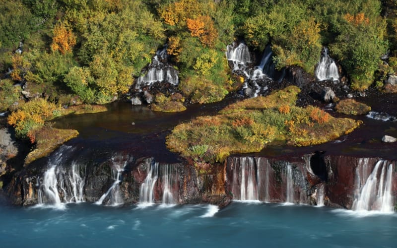 Aerial view of Hraunfossar waterfalls in autumn, with multiple cascades emerging from rust-colored and black lava rock into a vivid turquois