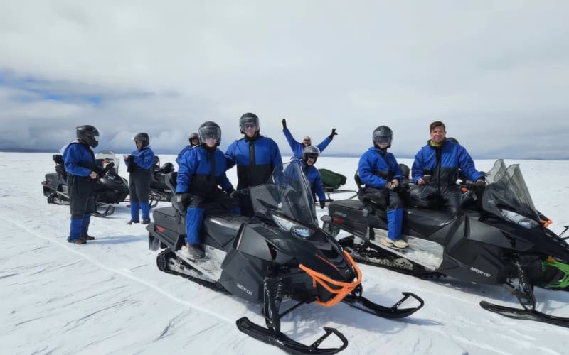 Group of eight visitors in matching blue snowmobile suits and helmets posing with Arctic Cat snowmobiles on the flat white surface of Langjo