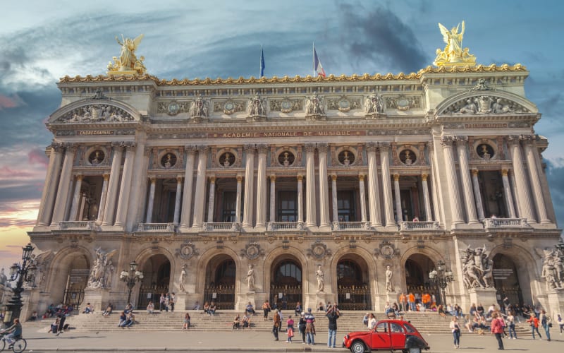 Ornate façade of the Opéra Garnier in Paris, with gilded rooftop sculptures, pedestrians and a red vintage car.