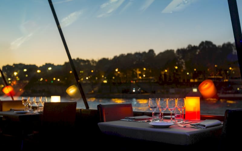 Dinner table with wine glasses and a glowing orange lamp beside panoramic windows overlooking the Seine at twilight.