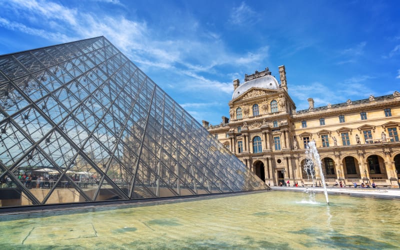 Louvre glass pyramid beside a shallow fountain pool, with the ornate museum façade beneath a bright blue sky.