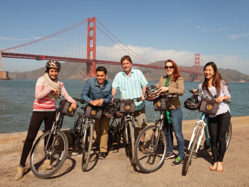 Group of five cyclists with bikes and backpacks posing on a beach with the Golden Gate Bridge in the background