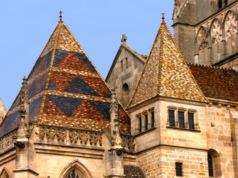 Close-up view of the colorful patterned tiled roofs and stone towers of Autun Cathedral in Burgundy, France, against a clear blue sky