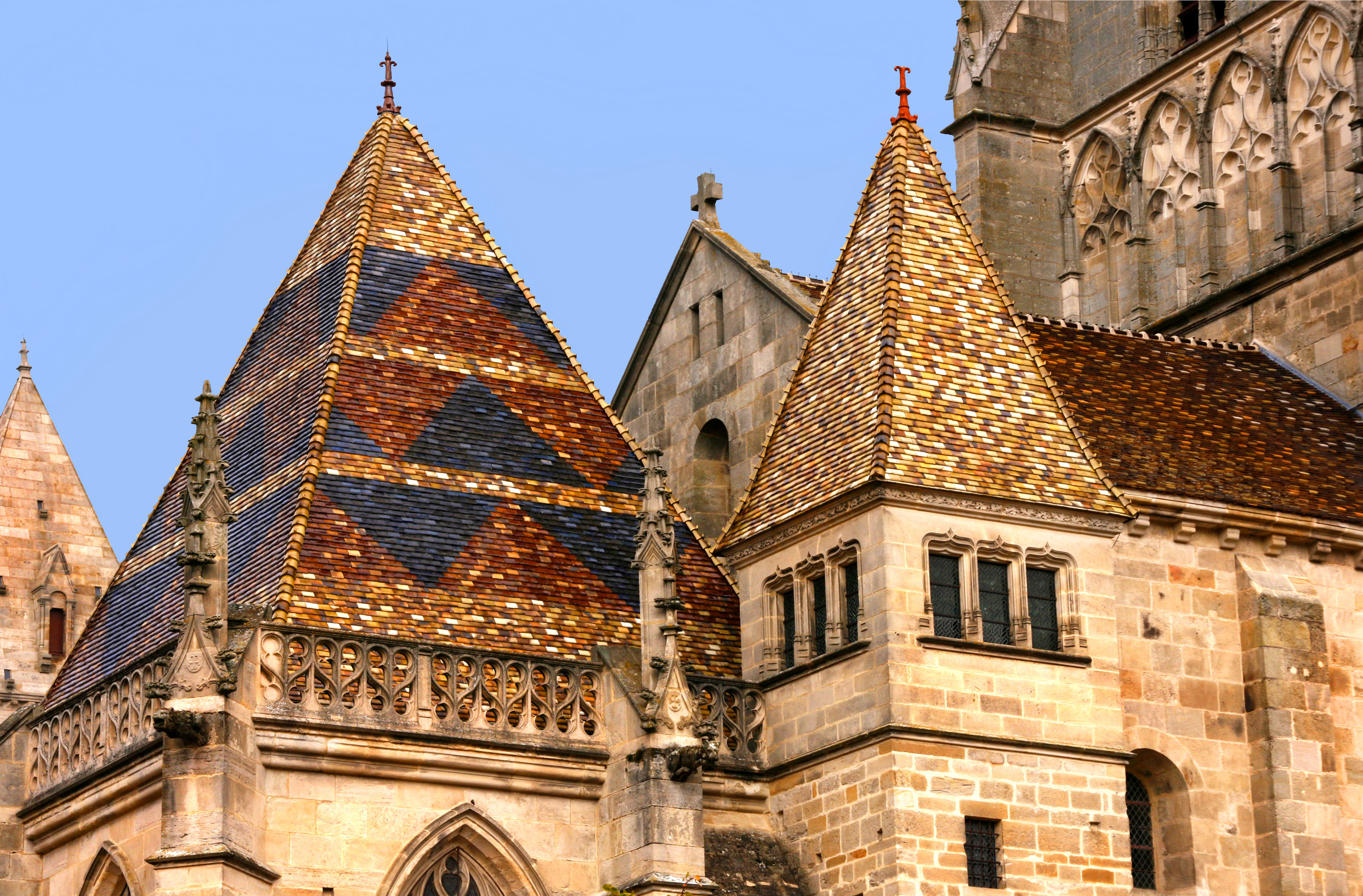 Close-up view of the colorful patterned tiled roofs and stone towers of Autun Cathedral in Burgundy, France, against a clear blue sky