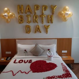 Romantic bedroom surprise with rose petals heart design, 'Happy Birthday' banner, and balloons.