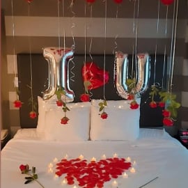 Romantic room decor with heart-shaped rose petals, red balloons, 'I❤ U' banner, cake, and candles.