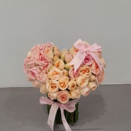 A heart-shaped arrangement of pink and peach roses, tied with a pink ribbon and resting in a clear vase.