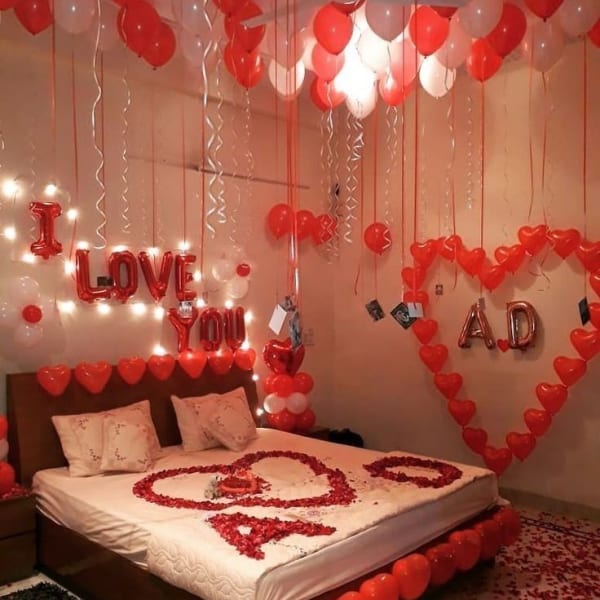 Romantic room decor featuring red rose petals, heart balloons, 'I LOVE YOU' banner, and LED lights.