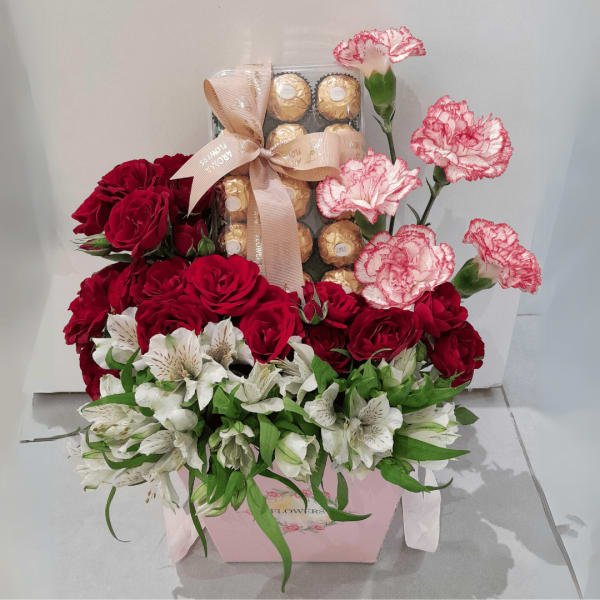 A vibrant flower box featuring red roses, pink alstroemeria, carnations, and Ferrero Rocher chocolates.