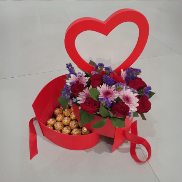 A flower box overflowing with red roses, purple chrysanthemums, blue statice, green ruscus, and Ferrero Rocher chocolates.