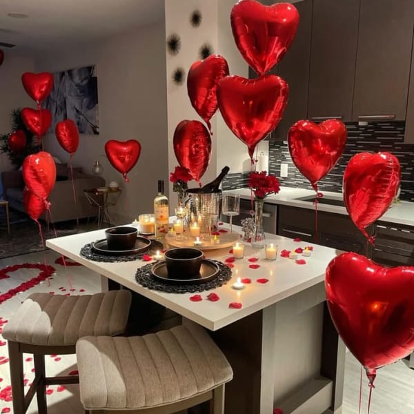 Romantic dinner setup with red heart balloons, rose petals forming hearts on the floor, scattered rose petals on the table and floor, and two red rose centerpieces with candles on a white table.