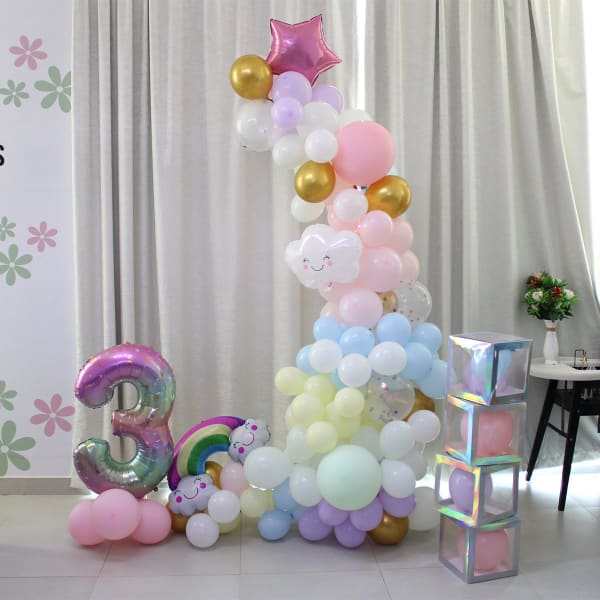 Whimsical unicorn birthday party decoration featuring a rainbow of pastel balloons, a large number three balloon, a cute unicorn, and stacked transparent balloon boxes.
