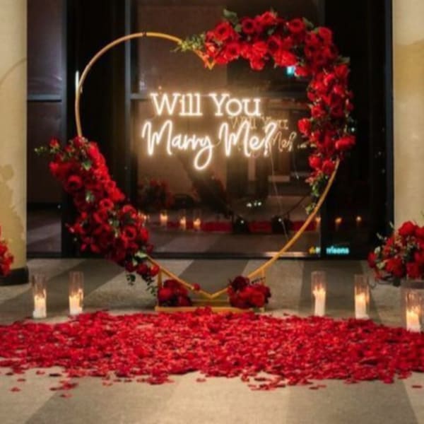 Romantic proposal setup with a red rose arch, neon sign, candles, and rose petals on the floor.