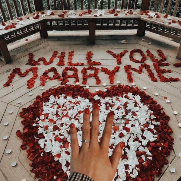 Romantic proposal scene with “Will You Marry Me” in rose petals, heart-shaped petal design, candles, and a hand with engagement ring.