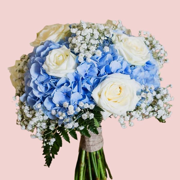 Bouquet with blue hydrangeas, white roses, baby’s breath, and greenery wrapped in rustic fabric.