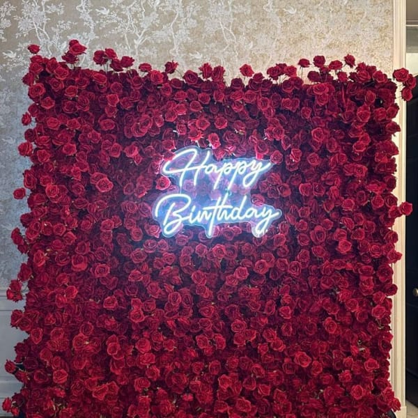A large square backdrop covered in deep red artificial roses with a glowing blue neon sign in the center that reads 