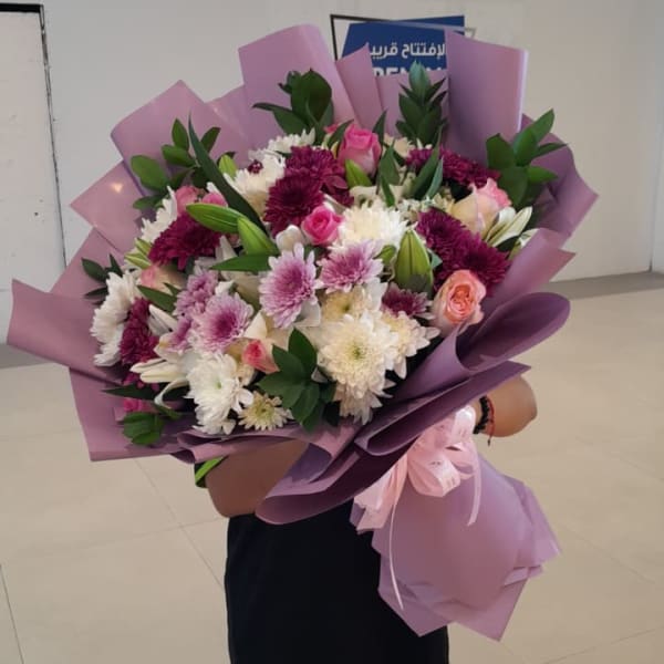 A person holding the large, beautifully arranged Floral Bliss Bouquet wrapped in purple paper with a pink ribbon.