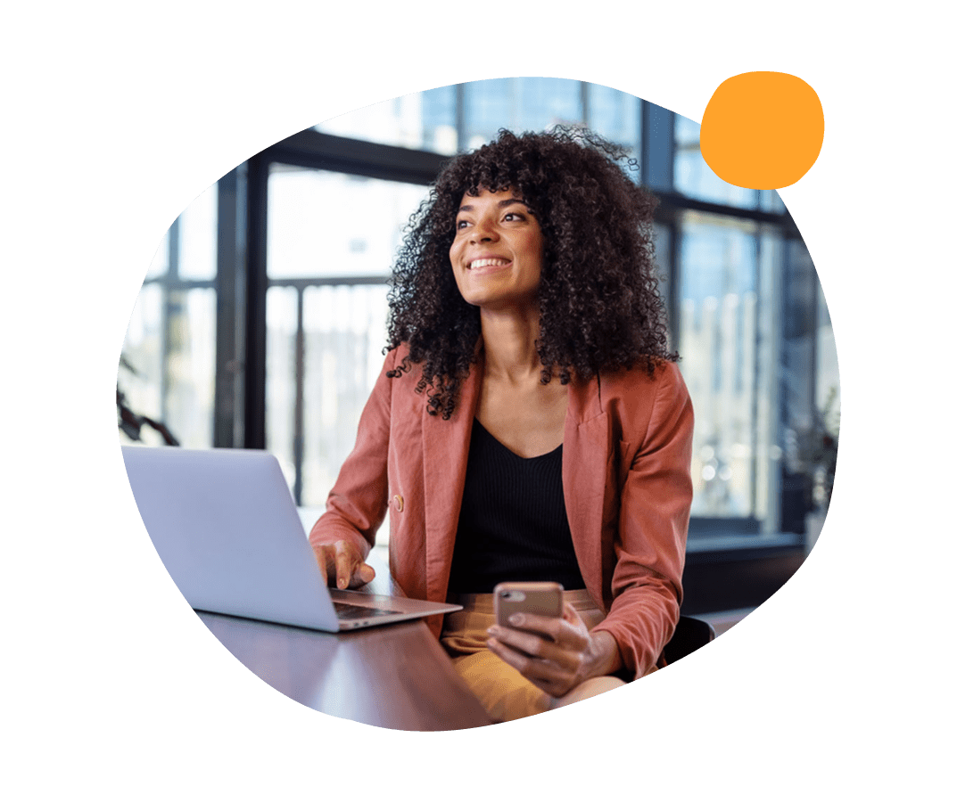 smiling woman using laptop and smartphone in modern office, curly hair and business attire in bright workspace