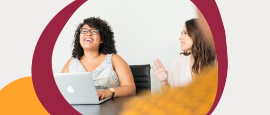 smiling women in a meeting, one with glasses, laptop on table, friendly interaction, modern office setting, happy coworkers collaborating in office