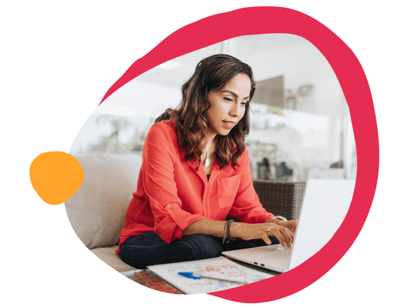 working woman in red shirt using laptop, focused expression, casual setting with notebook and pen on table, remote work lifestyle