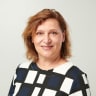 smiling woman with wavy brown hair wearing a geometric patterned blouse, neutral background in a professional portrait setting