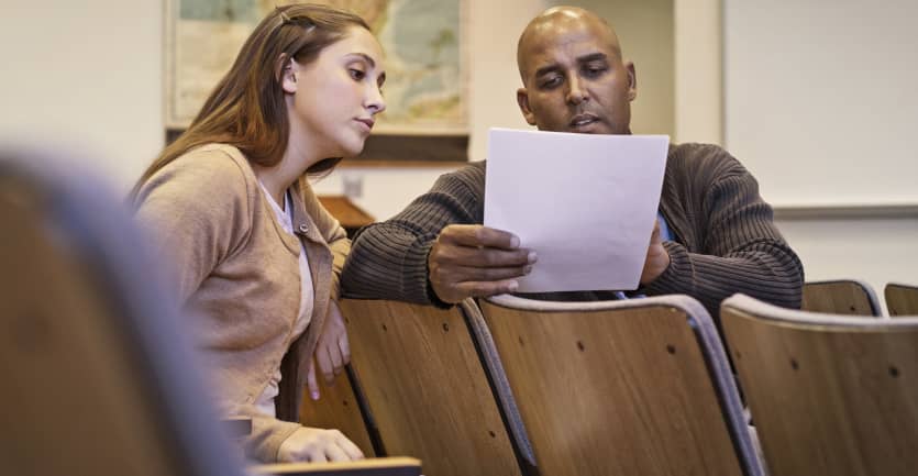 A student casually consults her professor about a printed essay, which they discuss in an empty lecture hall.