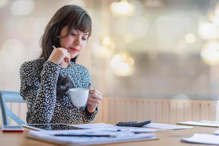 Woman drinking coffee while reading spreadsheets with a calculator