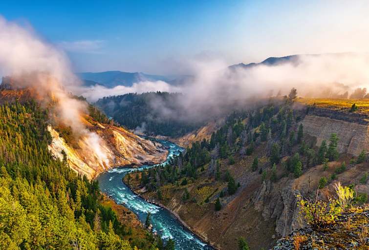 A wide angle summer morning view captured from the Calcite Springs View Area in Yellowstone National Park, Wyoming. Far below is the winding Yellowstone River, with the steaming Calcite Springs on the left bank of the river. Tall, tree-lined cliffs rise steeply on either side of the river and into the misty morning.