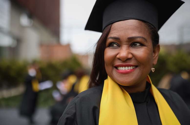 Black female doctoral graduate smiling at her commencement ceremony. She is wearing a black cap and gown and a yellow sash.